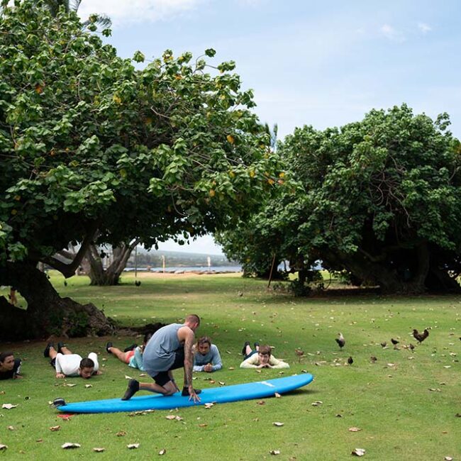 A man lies on a surfboard in a park, surrounded by grass and trees, enjoying a sunny day outdoors.
